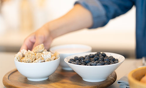 hand grabbing bowl of wheat cereal with a bowl of blueberries next to it