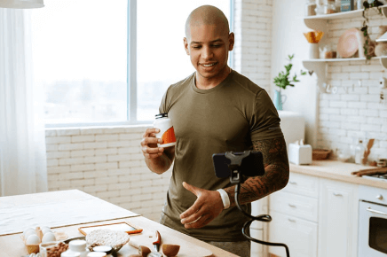 Male with tattoo sleeve talking to phone on tripod while holding bottle of supplements