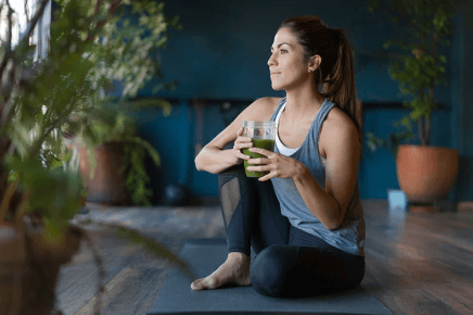 Female sitting on yoga mat holding cup of a green smoothie