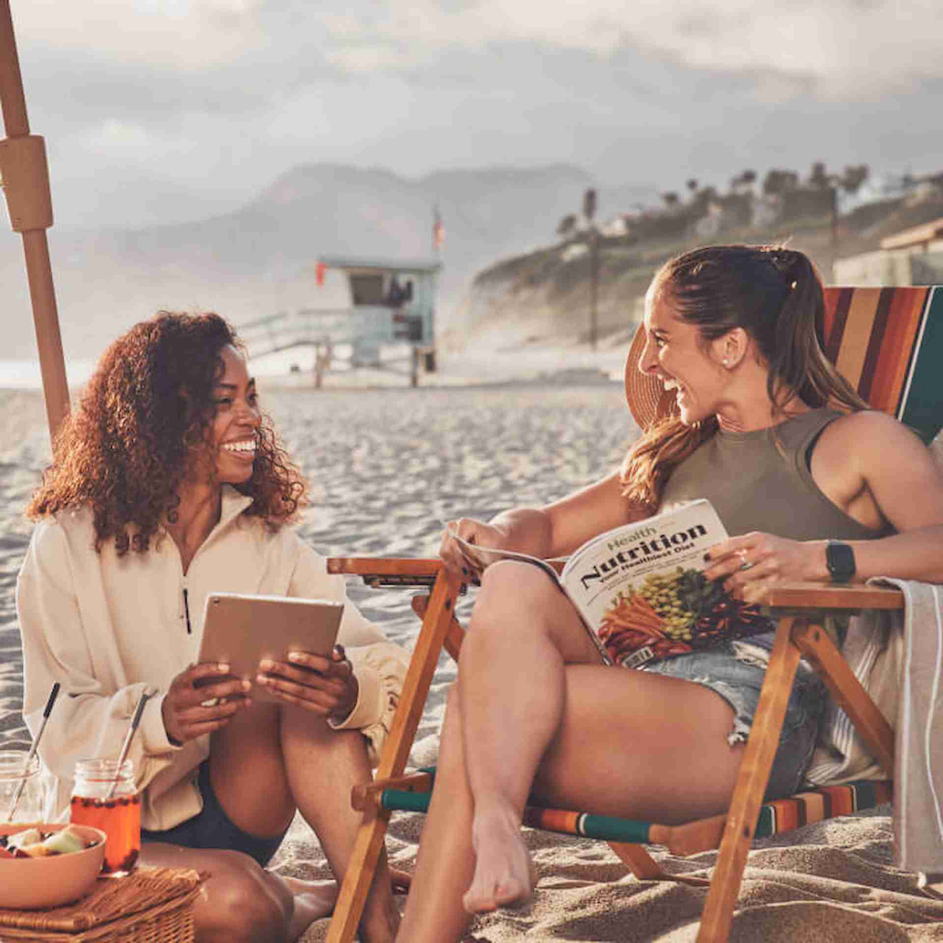 Two females sitting on the beach talking one holding magazine and other holding tablet