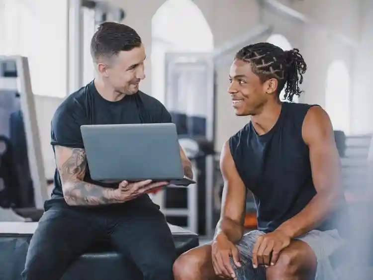 Two men in athletic wear sit in a gym; one holds a laptop and shows it to the other, during a NASM career consultation, as they both appear to be having a friendly conversation.